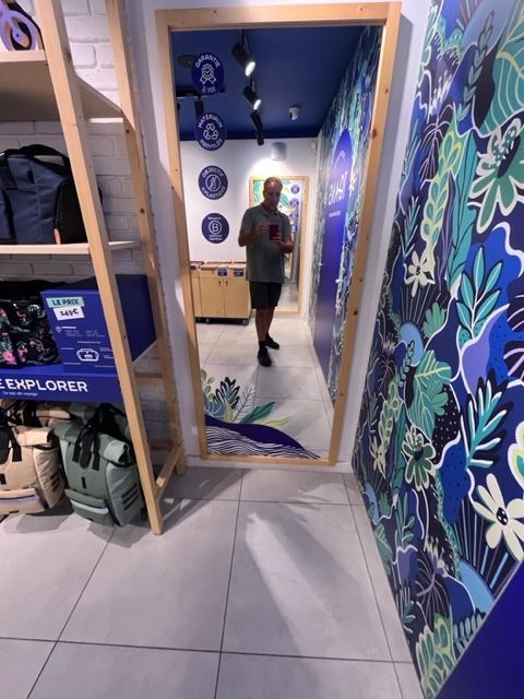Une personne prend un selfie devant un miroir d'un magasin, avec une fresque murale florale bleue.