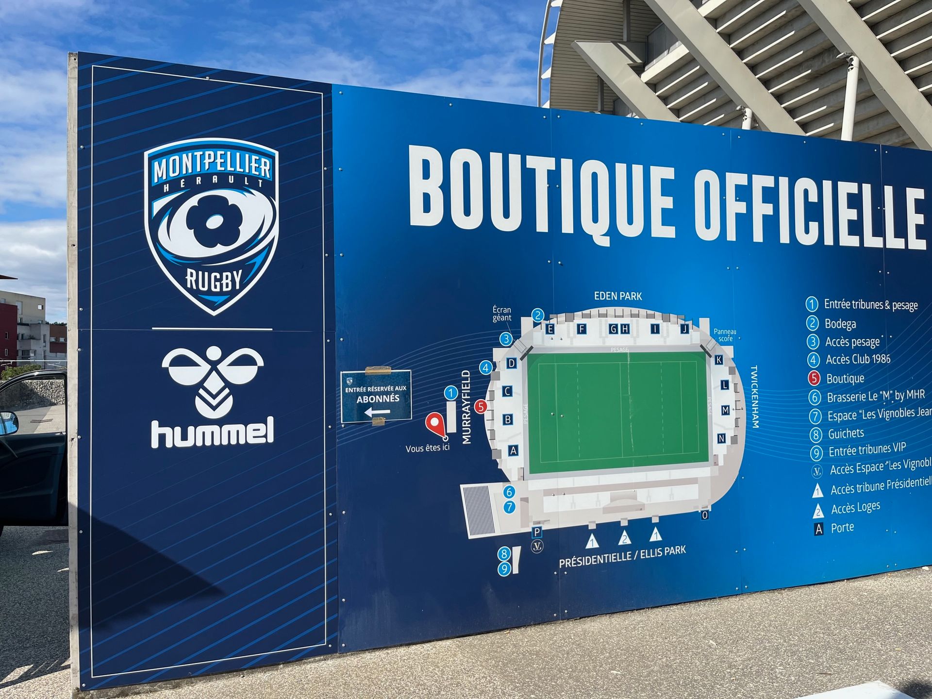 Panneau publicitaire bleu de la boutique officielle du Montpellier HSC avec le logo du club, la marque Hummel et le plan du stade.