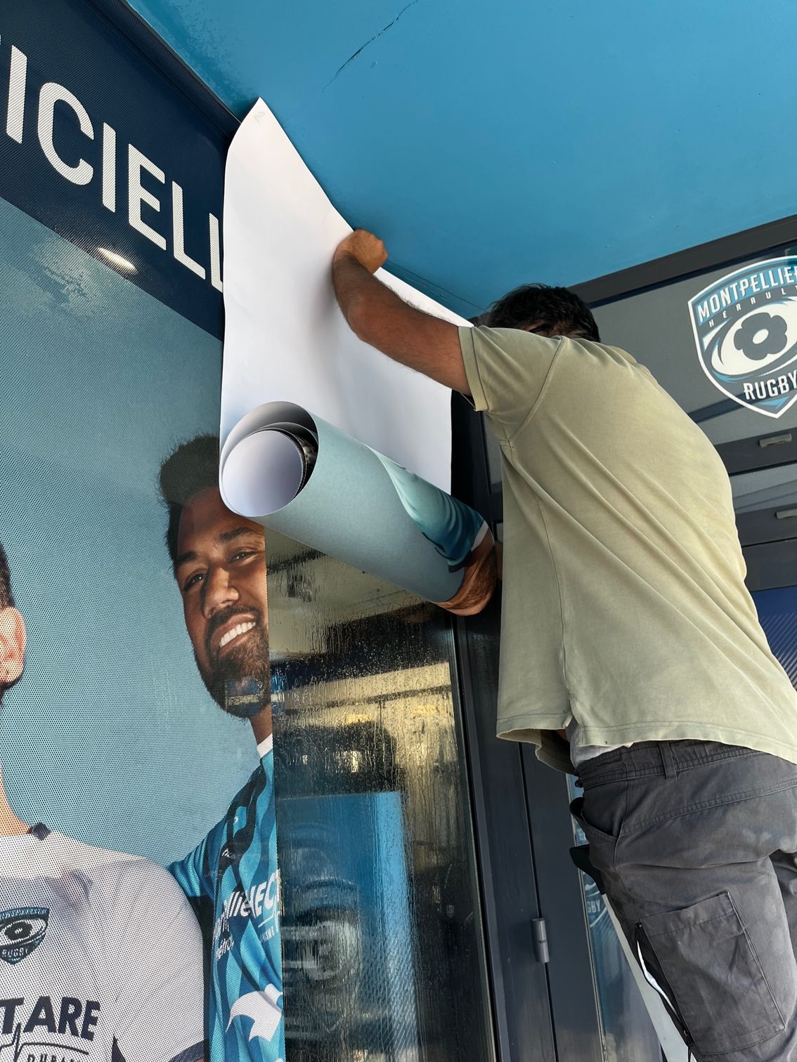 Un homme installe une grande affiche en vinyle sur un mur à côté d'une banderole publicitaire bleue.