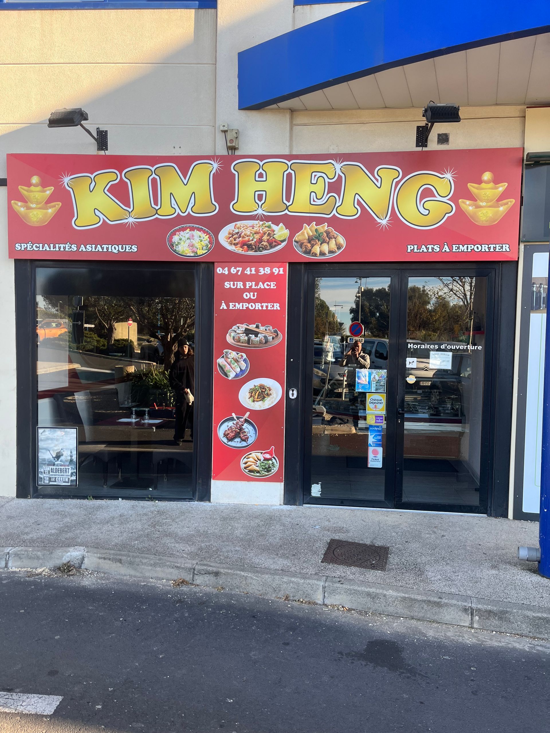 Façade avec une enseigne rouge Kim HENG au-dessus des portes vitrées et des images de nourriture sur la colonne d'entrée.