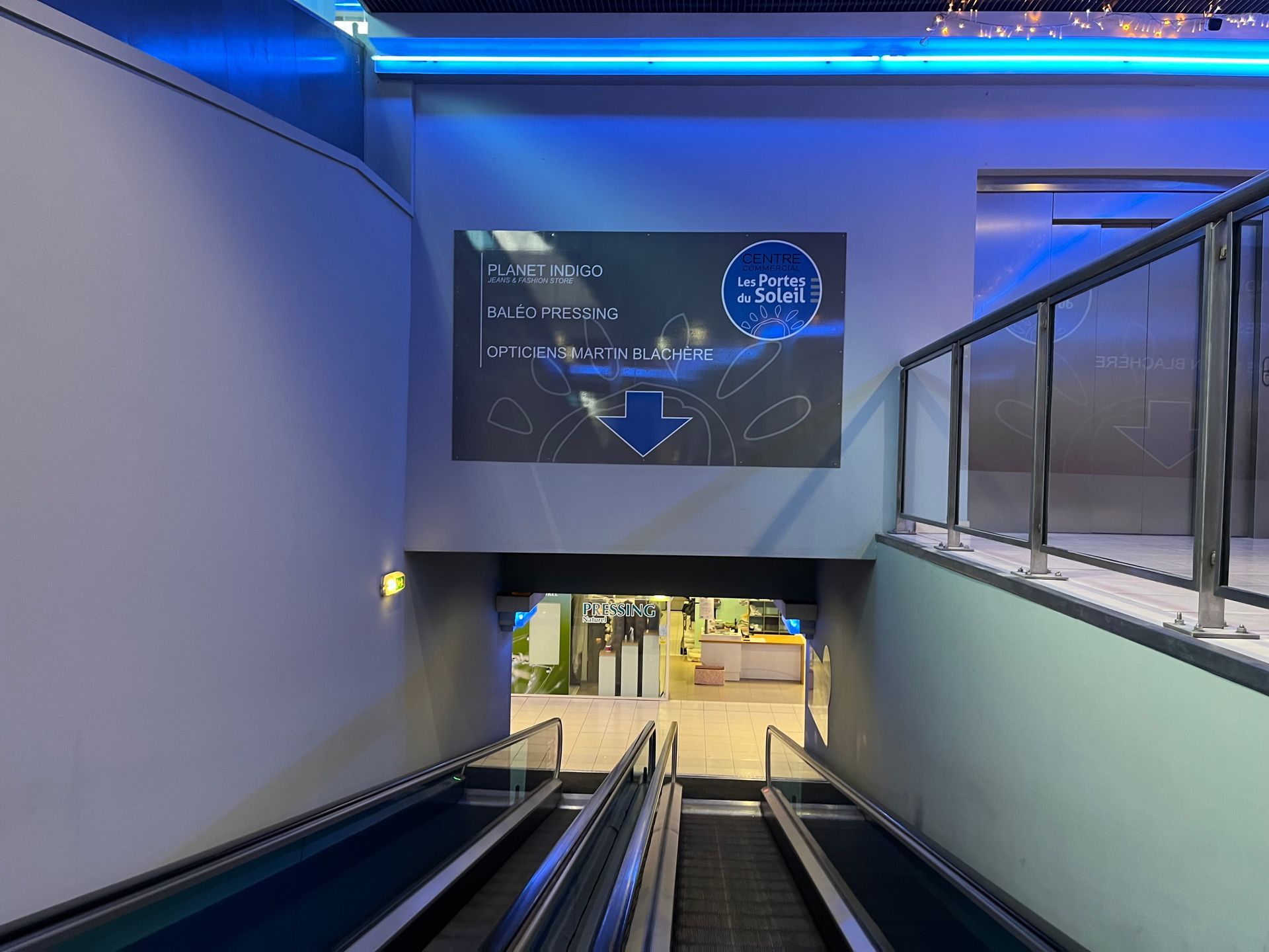 Escalator descendant vers un hall moderne éclairé en bleu avec un grand écran numérique devant.