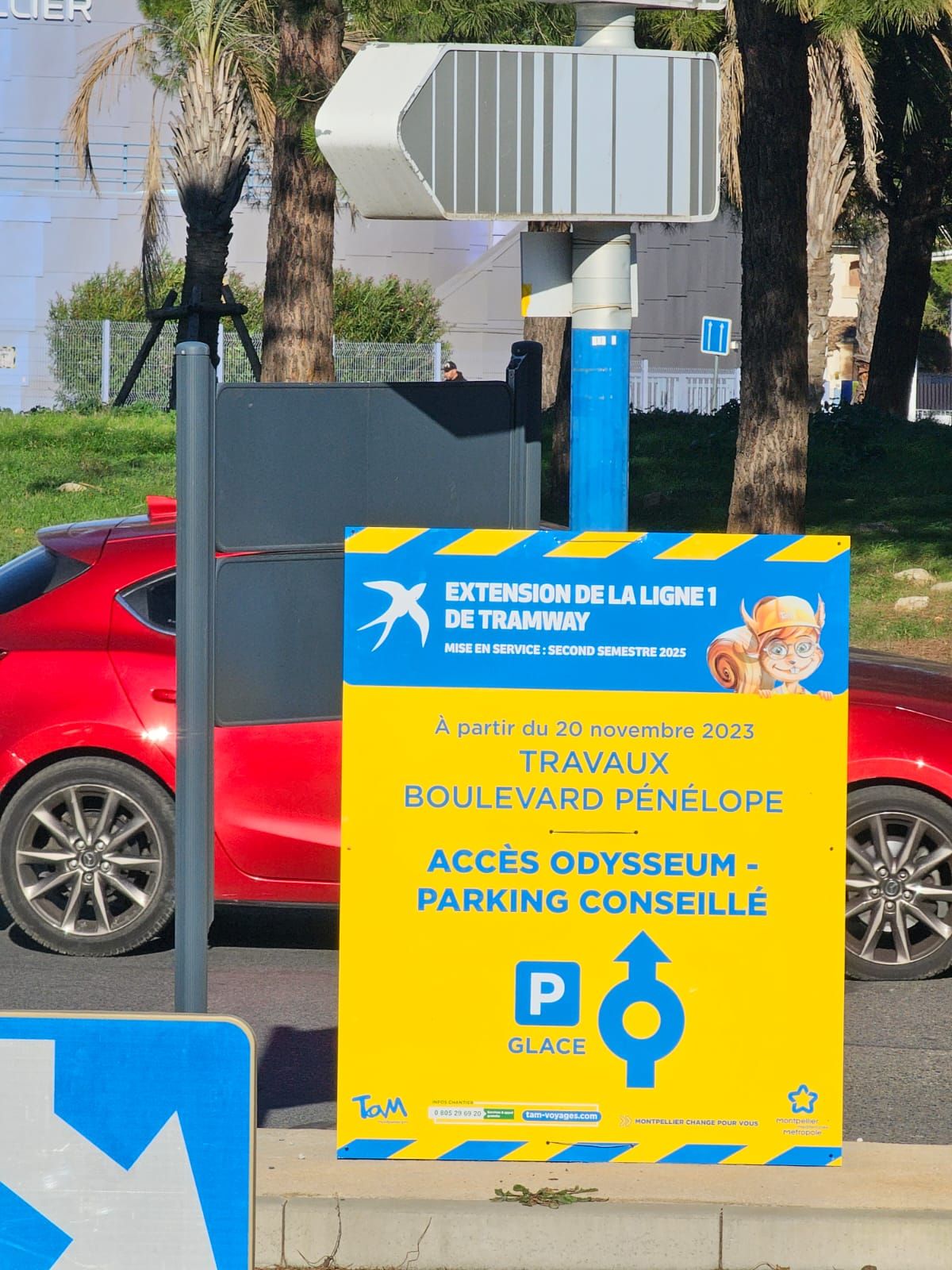 Panneau jaune de travaux signalant l'extension de la ligne de tramway, avec indication de stationnement accessible, à côté d'une voiture rouge et de palmiers.