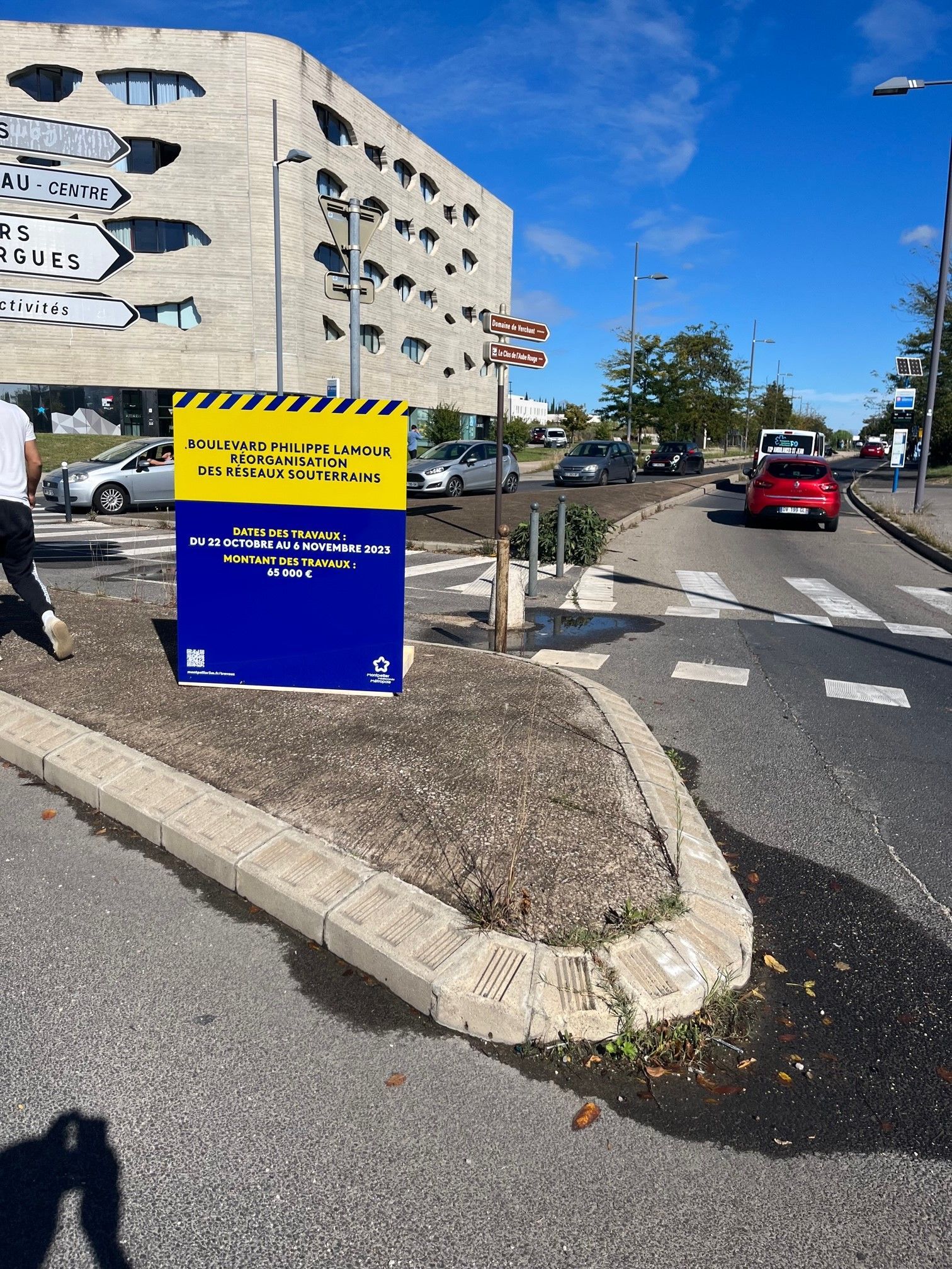 Panneau de travaux routiers sur un îlot de circulation en bordure de rue, avec des voitures, un parking et un ciel bleu.