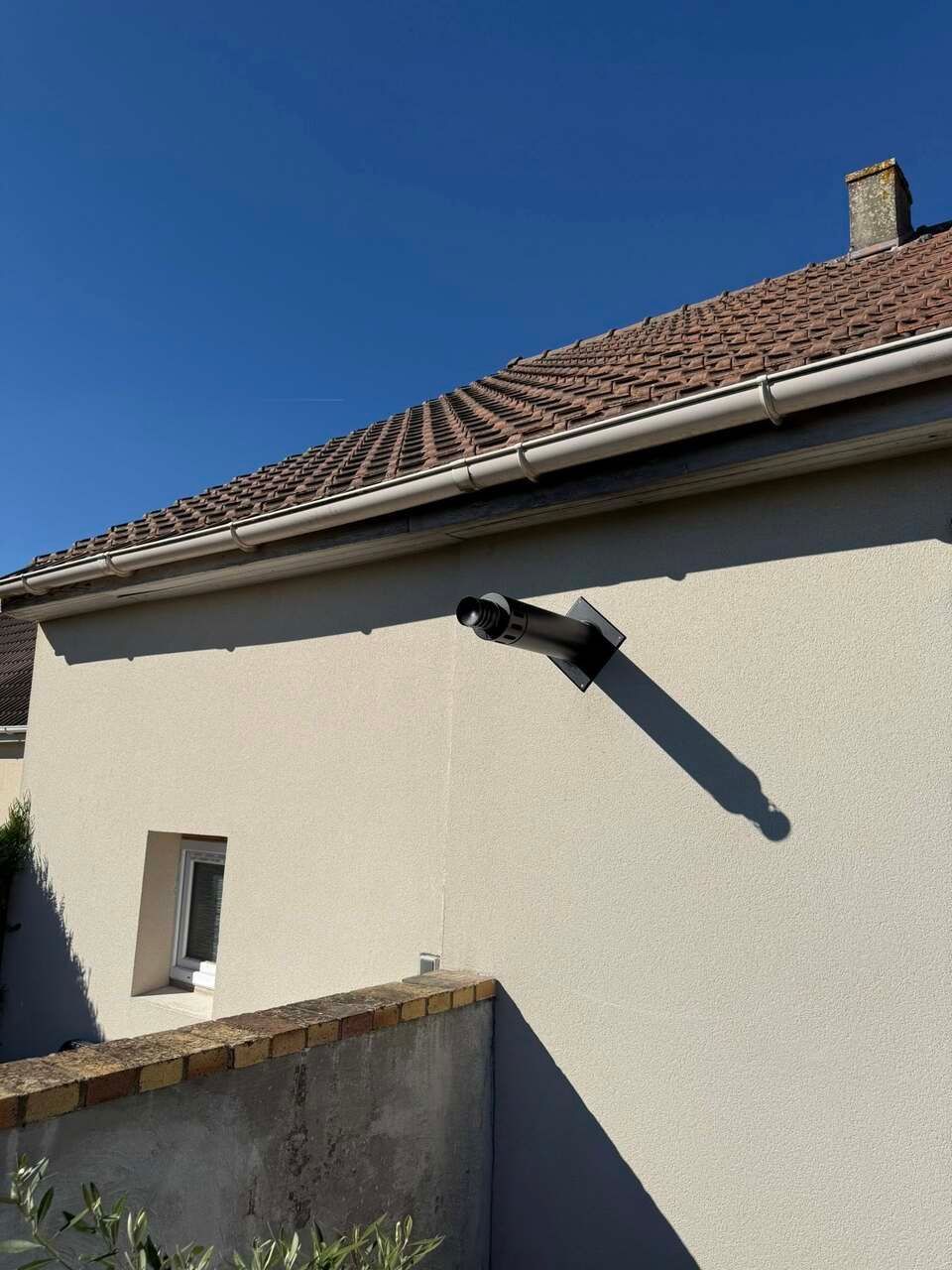 Extérieur d'un bâtiment avec un tuyau noir, une fenêtre et un toit sur fond de ciel bleu clair.