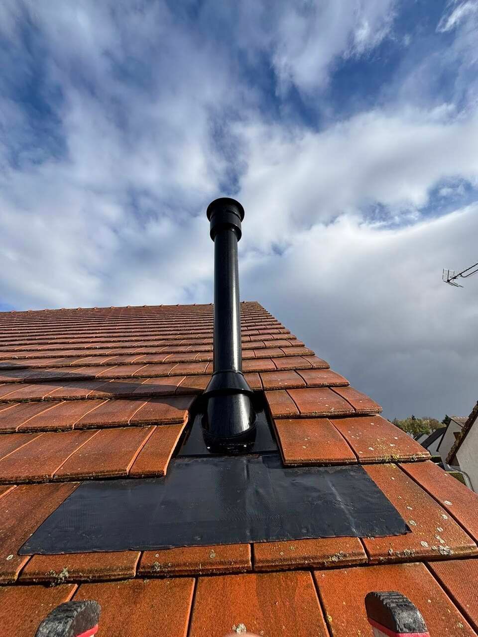 Tuyau noir d'évacuation des fumées sur un toit de tuiles, se détachant sur un ciel bleu nuageux.