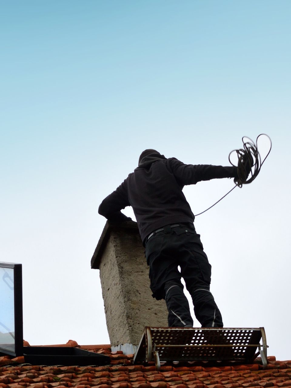 Un professionnel sur un toit, penché au-dessus d'une cheminée, tenant un câble, sur fond de ciel nuageux.