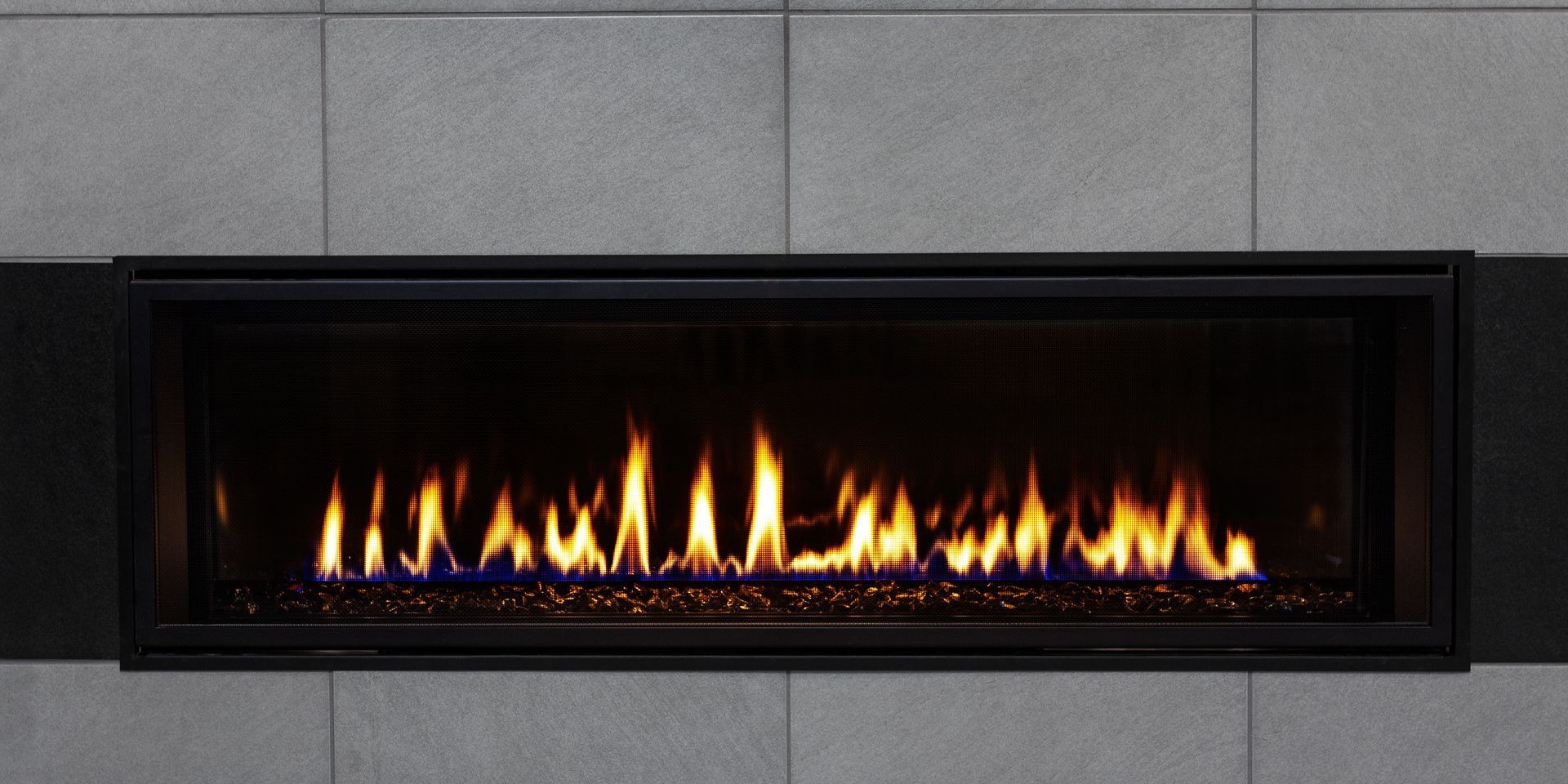 Une cheminée à gaz rectangulaire avec des flammes, encastrée dans un mur carrelé gris.