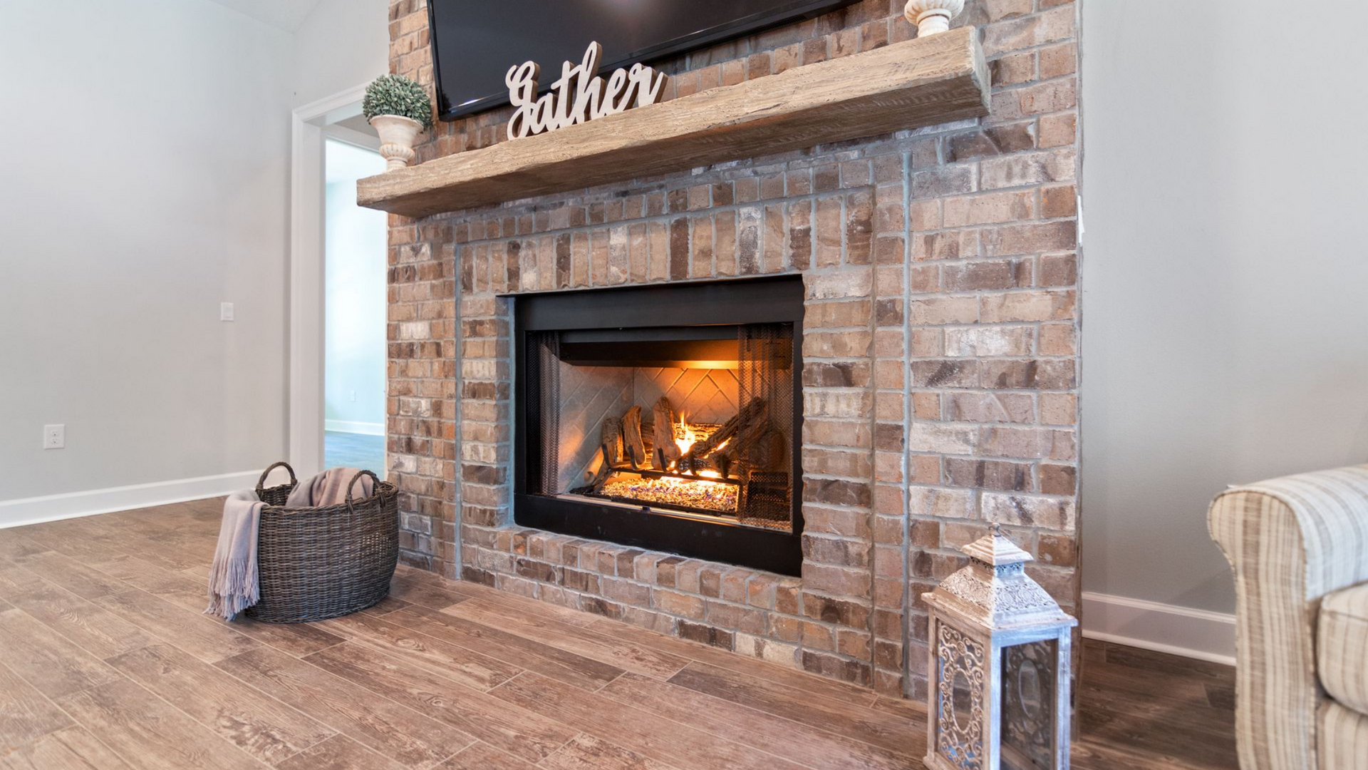 Cheminée avec entourage en briques, manteau en bois, feu qui crépite, télévision au-dessus. Panier en osier, lanterne et parquet.