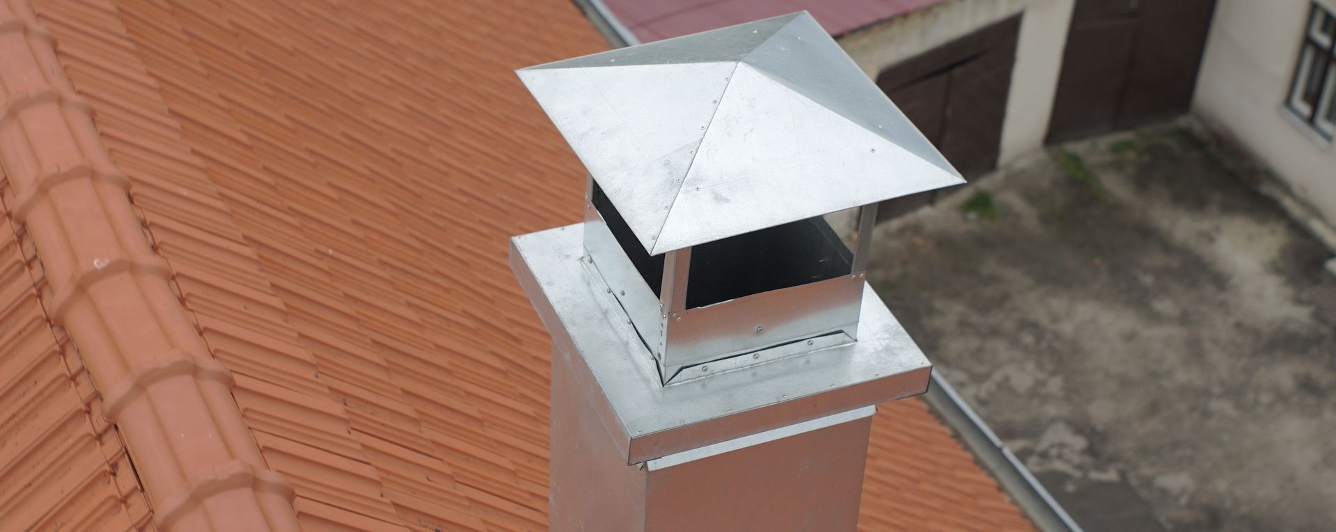 Cheminée à chapeau métallique sur un toit de tuiles rouges.