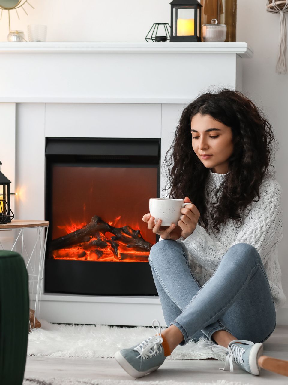 Femme assise près de la cheminée, tenant une tasse, vêtue d'un pull blanc et d'un jean.