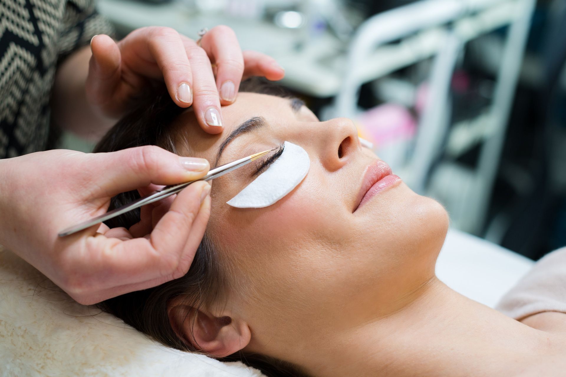 Mujer recibiendo extensiones de pestañas; técnico usando pinzas, ojo cubierto con una almohadilla.