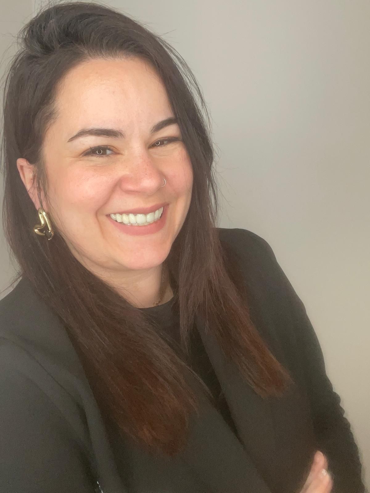 Mujer con cabello castaño y blazer oscuro sonríe ampliamente, con los brazos cruzados y pendientes de oro.