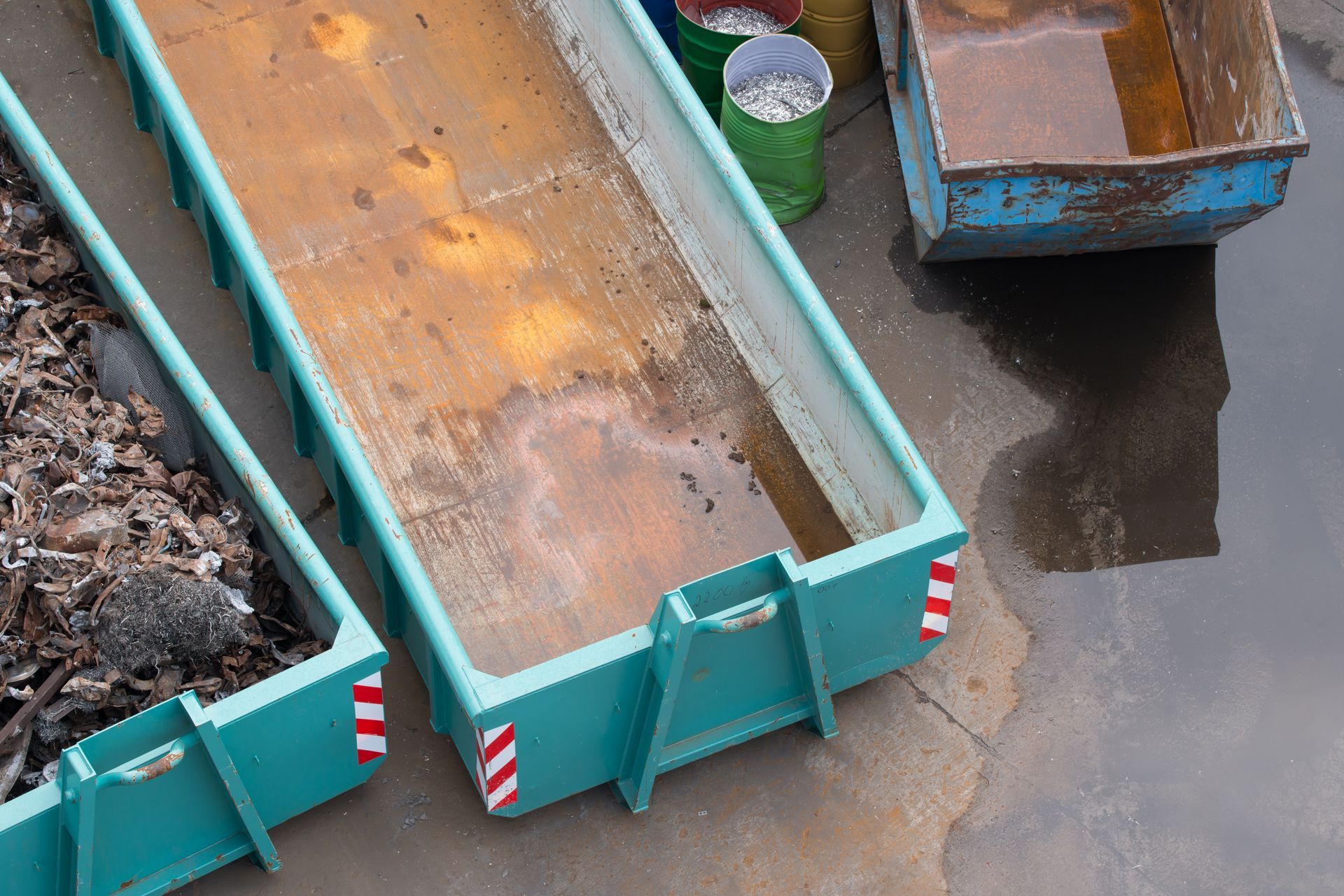Deux bennes métalliques turquoise, dont l'une contient des débris, sont posées sur une surface en béton à côté d'un conteneur