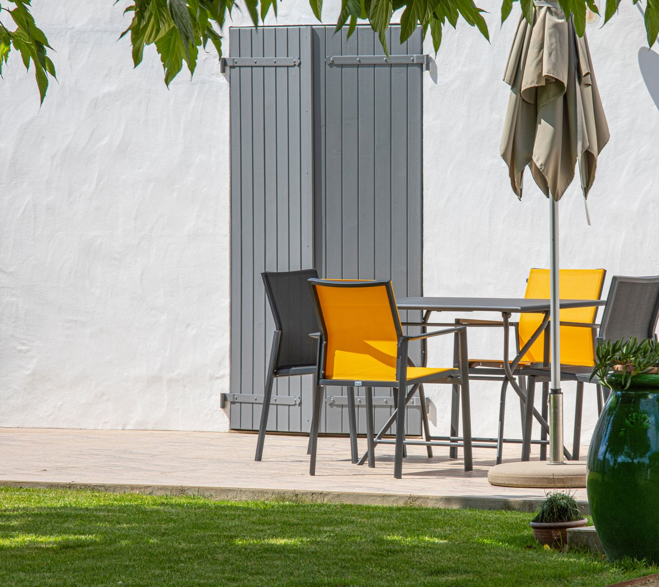 Terrasse extérieure avec table et chaises, volets gris et parasol. Chaises jaune et noire.