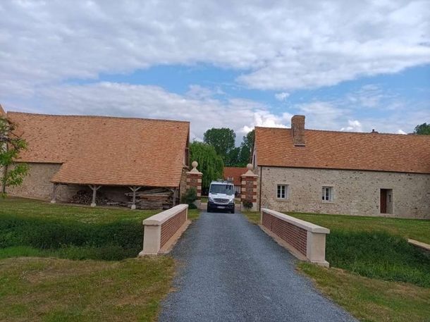 Fourgonnette sur une allée entre des bâtiments en pierre aux toits de tuiles rouges et un pont. Ciel nuageux.