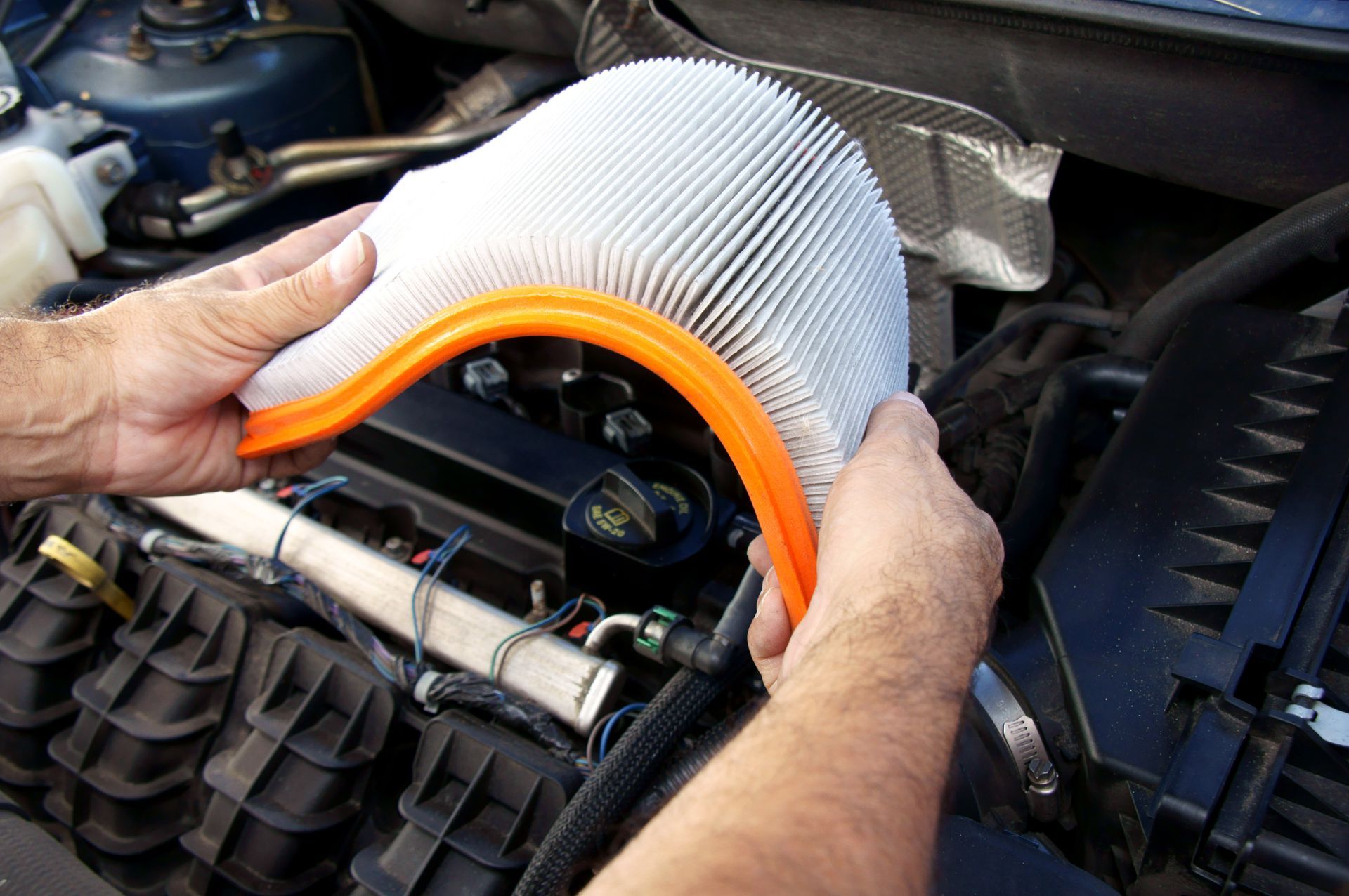 Des mains tiennent un filtre à air neuf au-dessus du compartiment moteur ; le filtre est blanc avec un cadre orange.