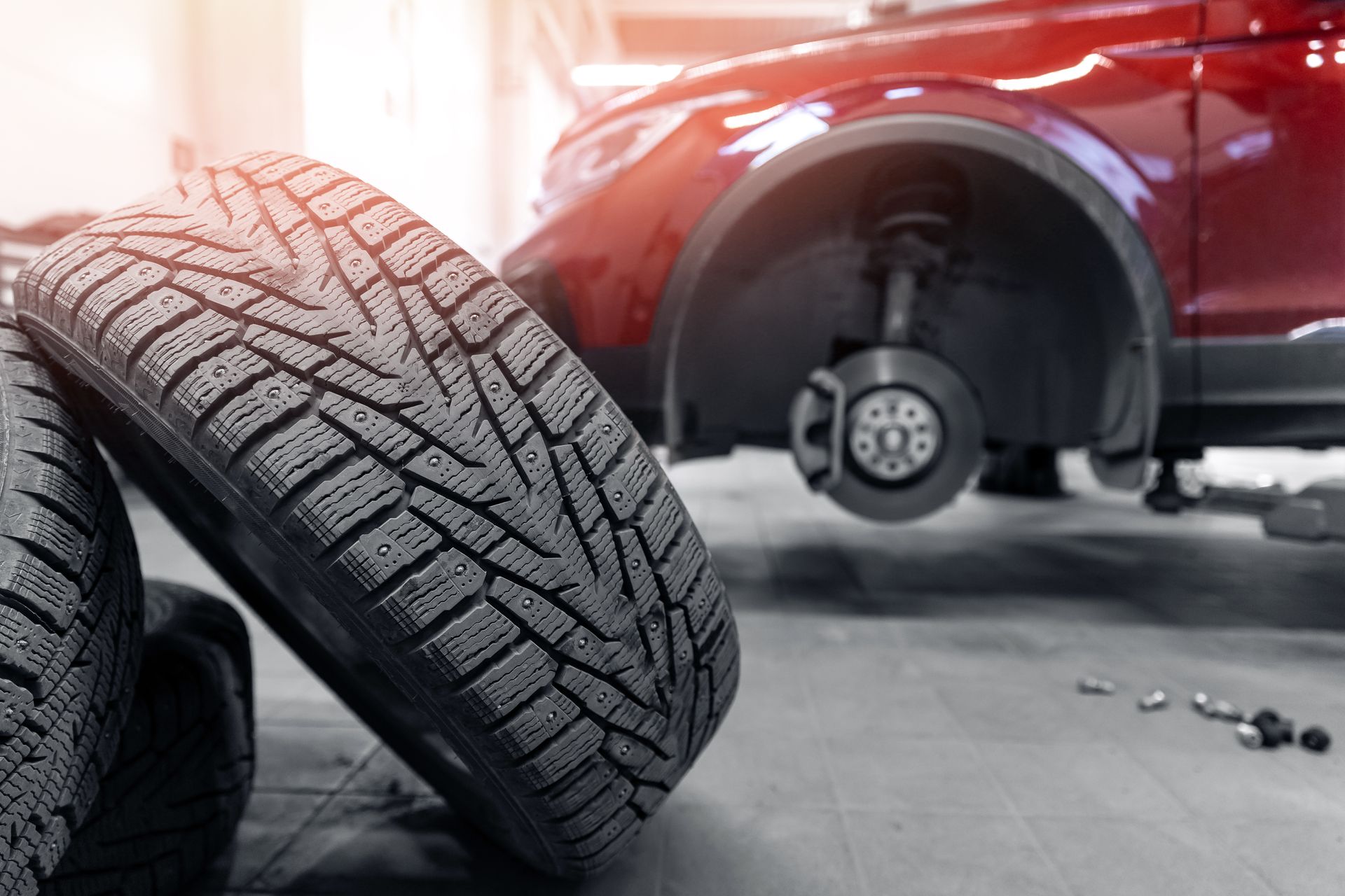 Deux pneus d'hiver appuyés contre une voiture rouge dont un pneu a été retiré, à l'intérieur d'un garage.