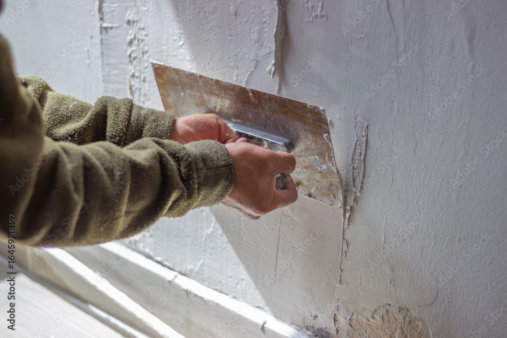 Eine Person, die eine Fleecejacke trägt, glättet mit einer Metallkelle Gips auf einer strukturierten Wand.