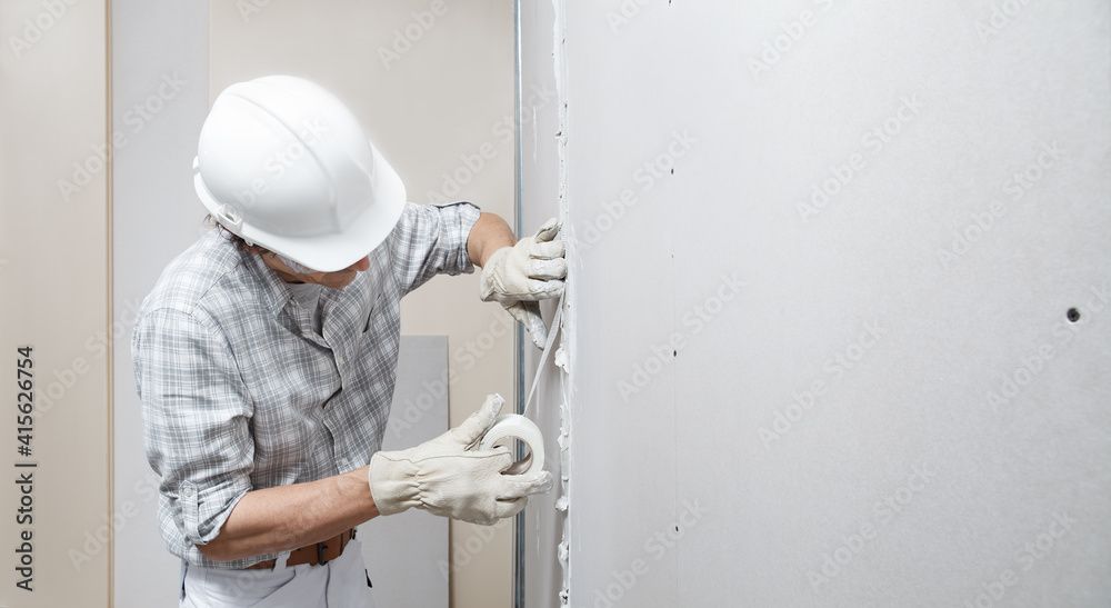 Ein Arbeiter mit Schutzhelm und Handschuhen bringt Fugenband an einer Gipskartonfuge an.
