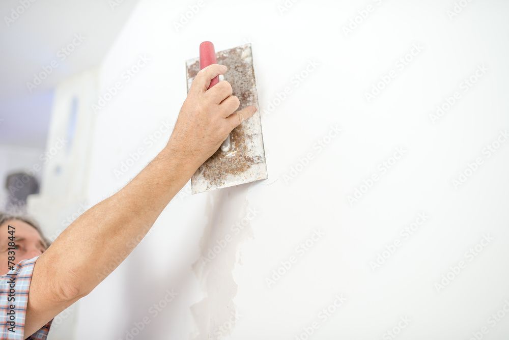 Eine Person benutzt eine Metallkelle, um Gips auf eine weisse Wand aufzutragen und zu glätten.