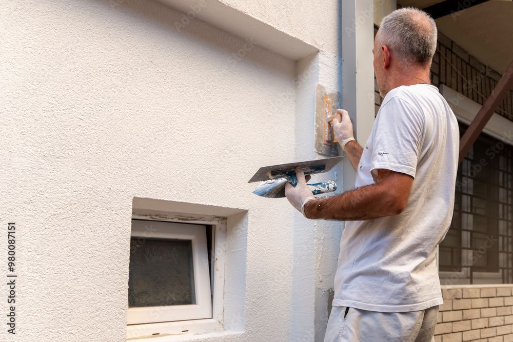 Eine Person in einem weissen Hemd glättet mit einer Kelle weissen Putz auf eine strukturierte Aussenwand in der Nähe eines kleinen Fensters.