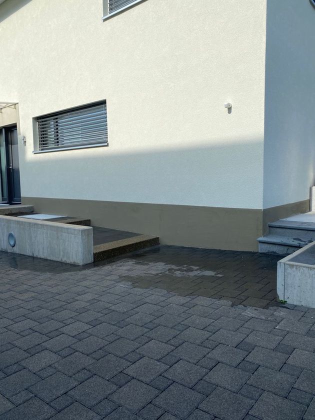 Eine Person in einem weissen Hemd glättet mit einer Kelle weissen Putz auf eine strukturierte Aussenwand in der Nähe eines kleinen Fensters.
