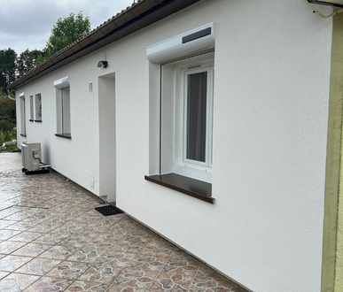 Maison blanche avec volets fermés, cadres de fenêtres sombres et patio pavé.