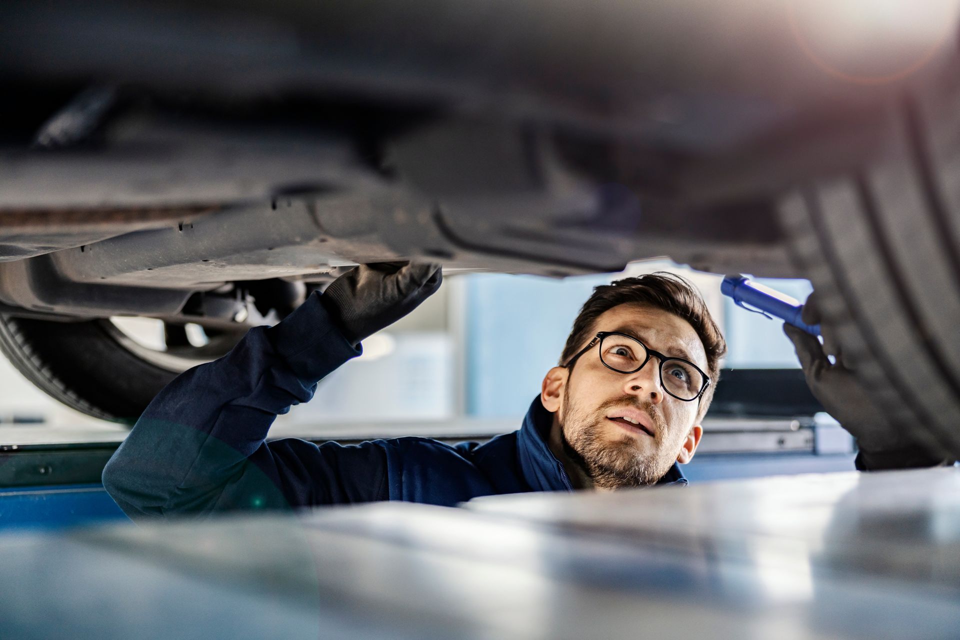 Un technicien portant des lunettes inspecte le dessous d'une voiture avec une lampe torche.
