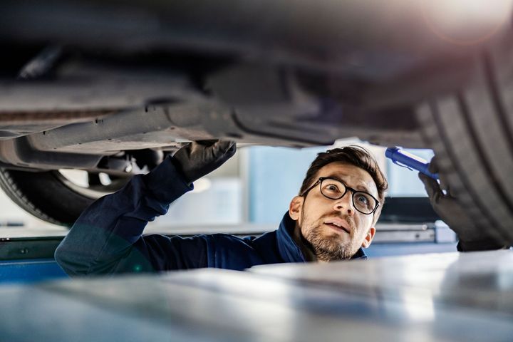 Un technicien portant des lunettes inspecte le dessous d'une voiture avec une lampe torche.