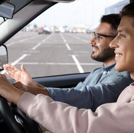 Dos hombres van en un coche; uno gesticula mientras hablan y, delante, se ve una pista de aeropuerto