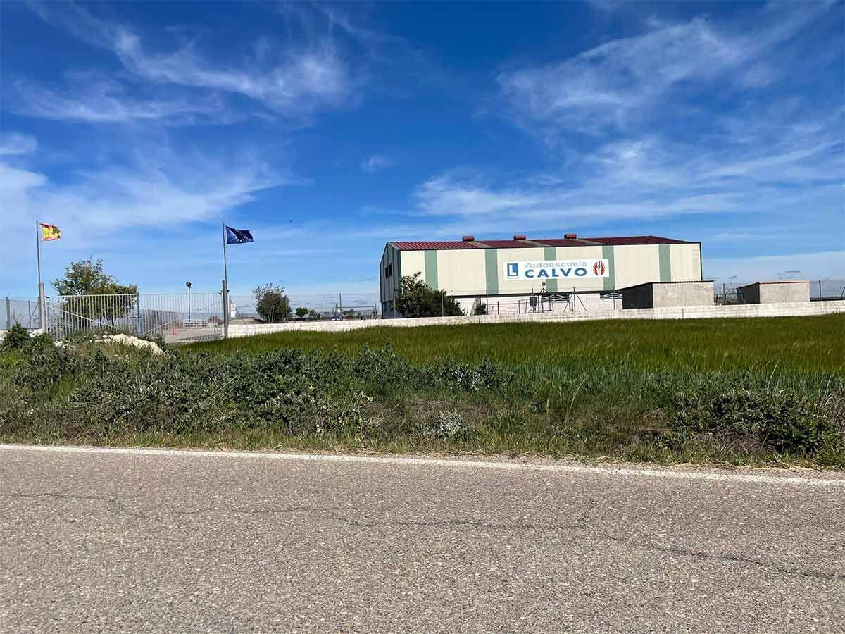 Una carretera ancha junto a un campo verde y un edificio grande bajo un cielo azul con banderas ondeando.