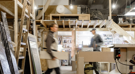 Des personnes travaillent le bois dans un atelier encombré. L'une d'elles passe avec une planche.