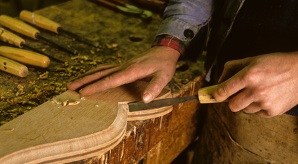 Une personne sculpte du bois au ciseau dans un atelier ; de la sciure est visible, des outils sont à proximité.