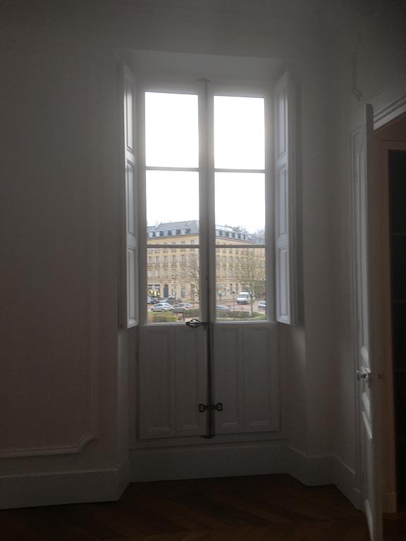 Fenêtre aux volets fermés donnant sur une rue et un immeuble. La pièce est baignée de lumière.