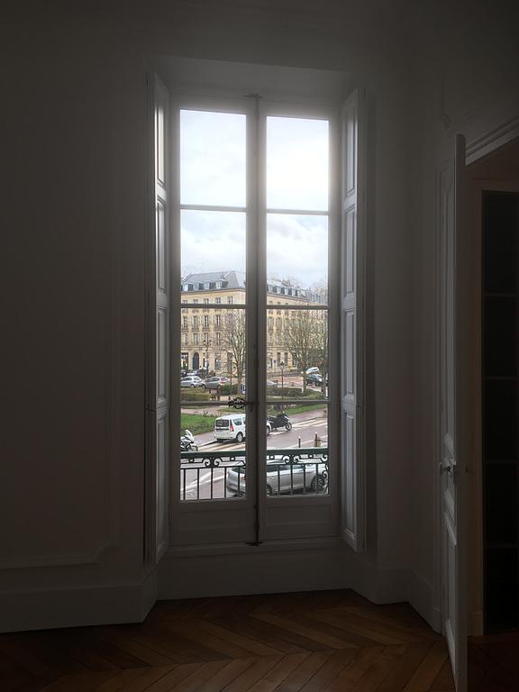 Une haute fenêtre à cadre blanc donne sur une rue de la ville, bordée de voitures et d'immeubles. La lumière du soleil inonde l'espace.