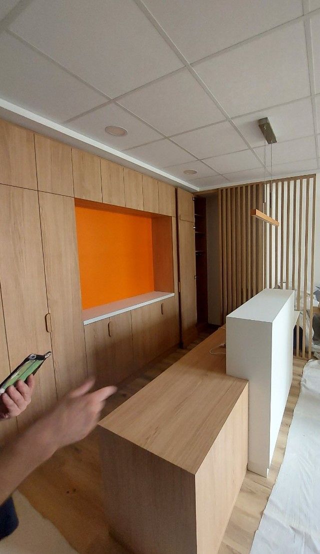 Intérieur d'une pièce avec des boiseries, une niche orange et un comptoir blanc et bois. Quelqu'un montre du doigt.
