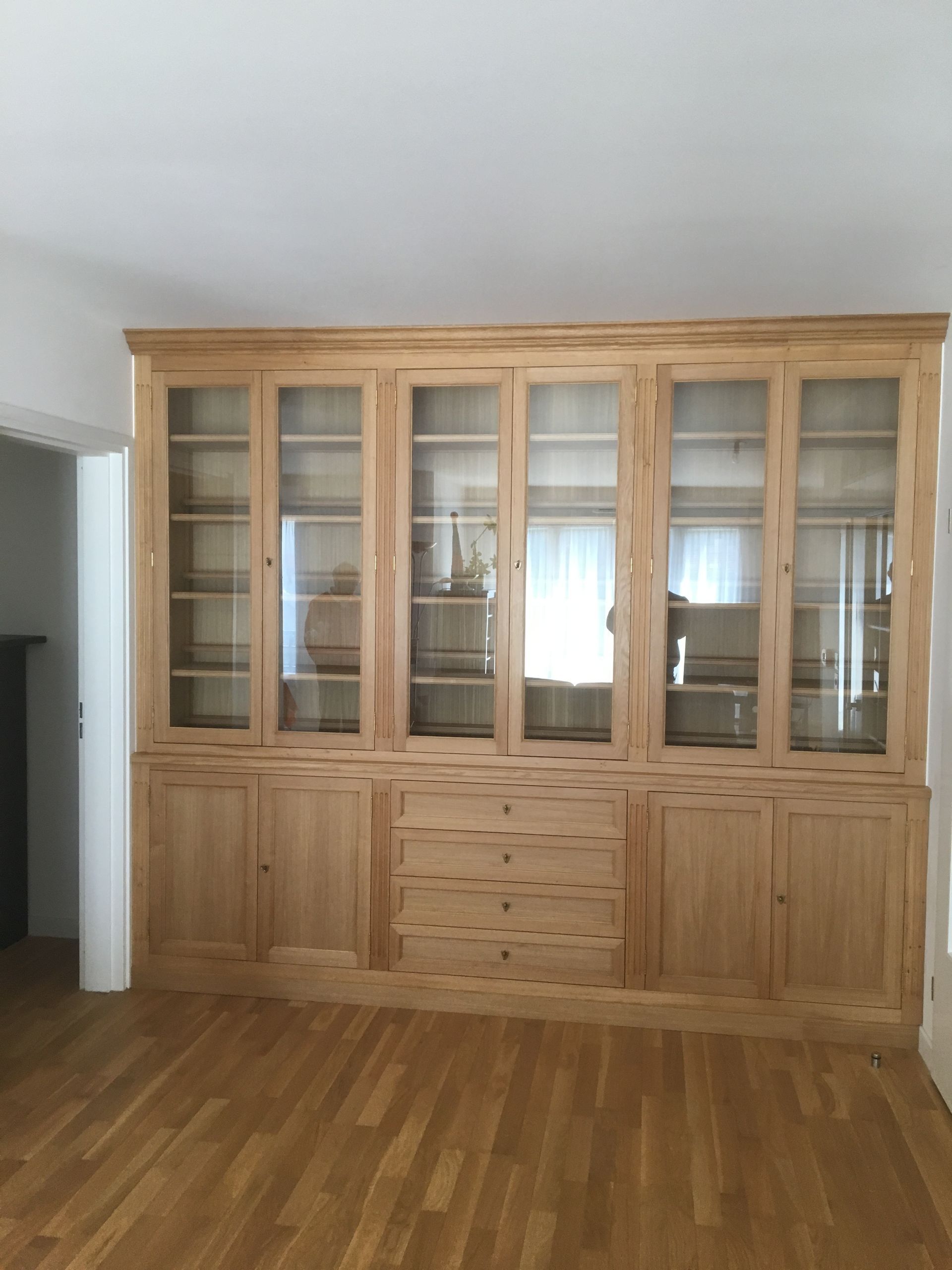 Meuble encastré en bois avec étagères vitrées, tiroirs et rangements inférieurs, dans une pièce avec parquet.