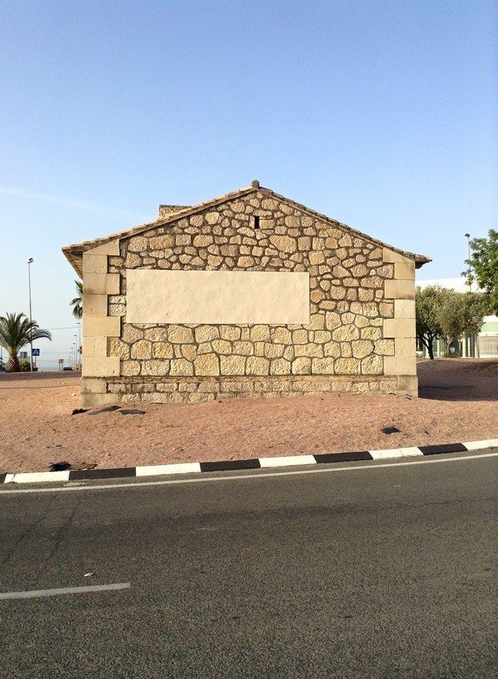 Edificio de piedra con un letrero rectangular de color claro en blanco, situado junto a una carretera pavimentada.