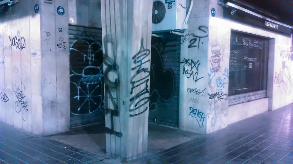 La esquina de un edificio por la noche, con contraventanas metálicas cubiertas de grafitis e iluminadas por una luz artificial fría.