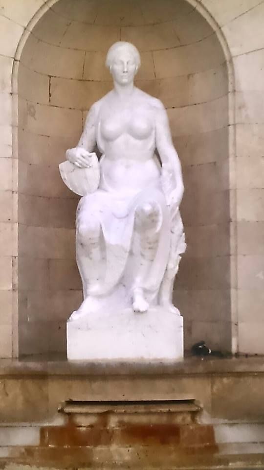 Una estatua de mármol blanco que representa una figura sentada sosteniendo un objeto, colocada dentro de una hornacina arqueada de piedra.