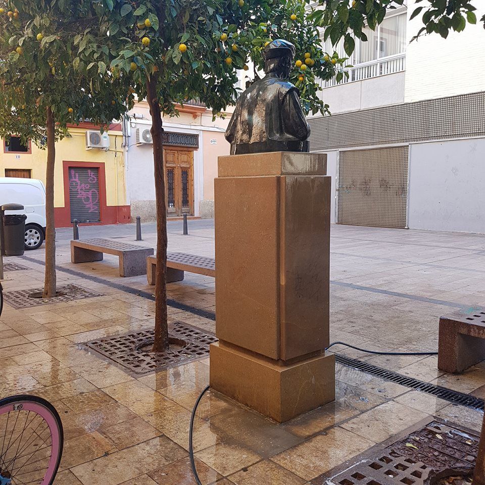 Un busto de bronce de una figura histórica sobre un pedestal de piedra en una plaza pública con naranjos y bancos de piedra.