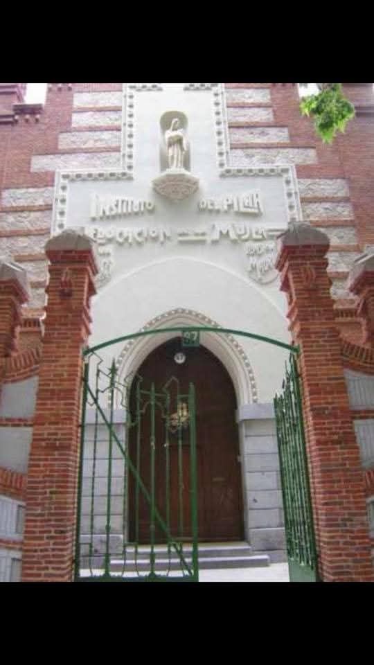 Fachada de ladrillo y piedra de un edificio con puerta de madera arqueada, verja de hierro y una estatua central en una pequeña hornacina.