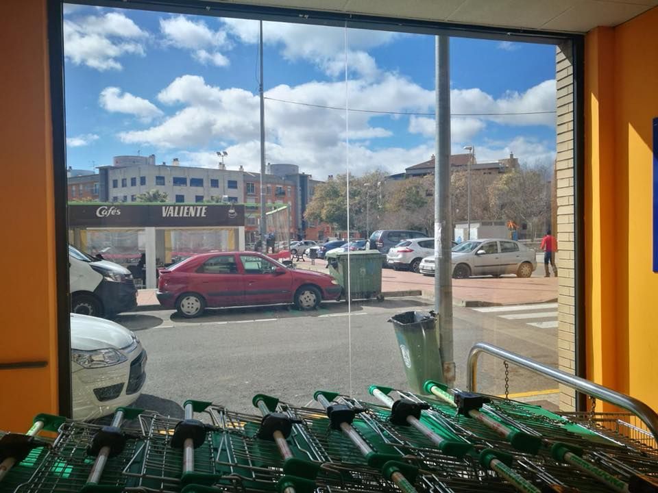 Vista a través del escaparate de una tienda que muestra un aparcamiento con varios coches, un contenedor de basura y carritos de la compra apilados en la parte delantera.