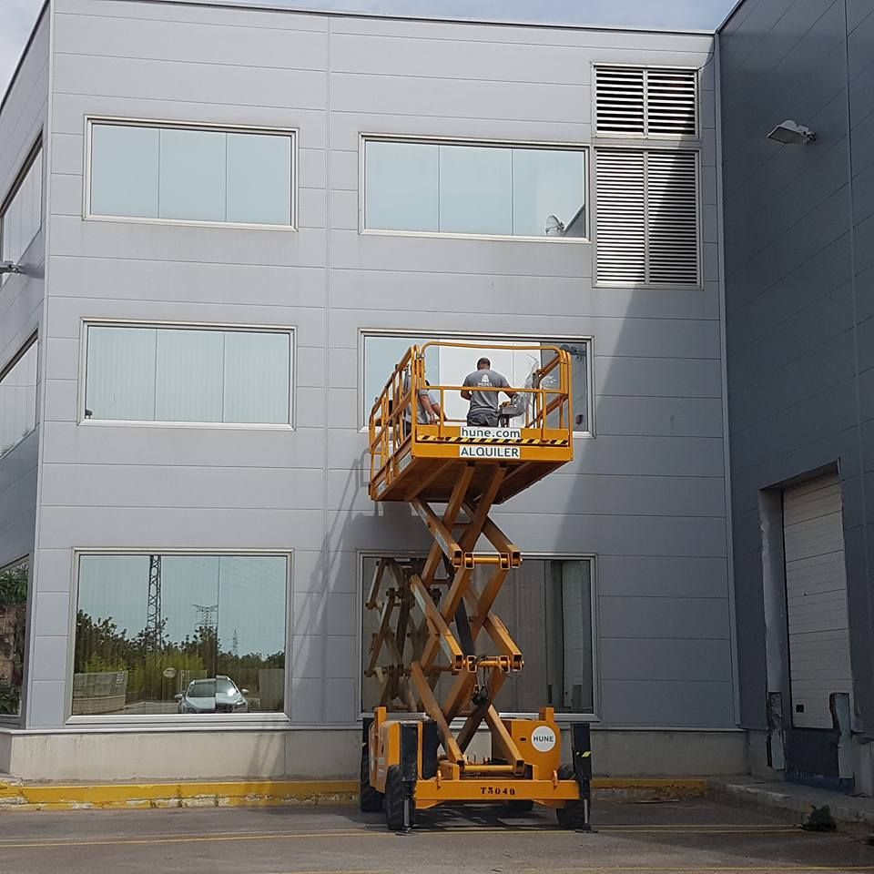 Un operario limpia las ventanas de la fachada de un edificio comercial de color gris claro utilizando una plataforma elevadora de tijera amarilla.