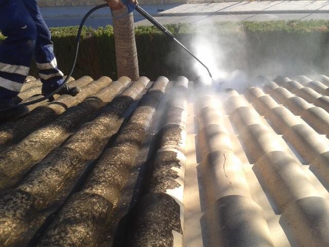 Una persona utiliza una hidrolimpiadora para limpiar las algas oscuras y la suciedad de las tejas curvas de color claro.