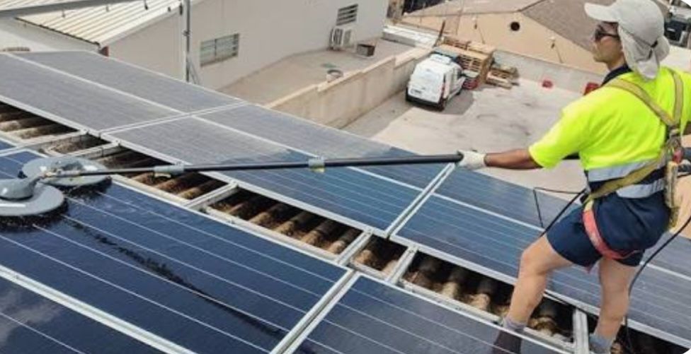 Un trabajador con una camisa de alta visibilidad utiliza un cepillo de mango largo para limpiar los paneles solares instalados en un tejado de tejas.