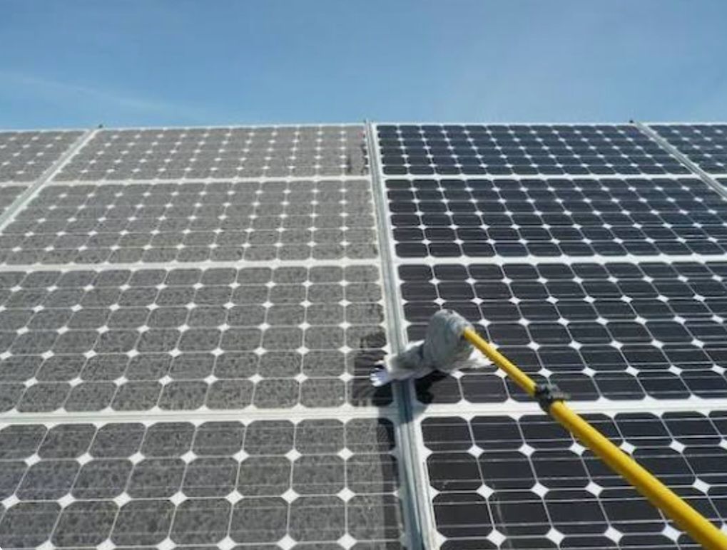 Un cepillo montado en un poste limpia un panel solar sucio, revelando una superficie más oscura e impoluta en comparación con el panel polvoriento que tiene al lado.