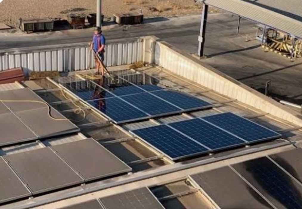 Una persona limpia una hilera de paneles solares oscuros en un tejado industrial plano con una herramienta de mango largo.