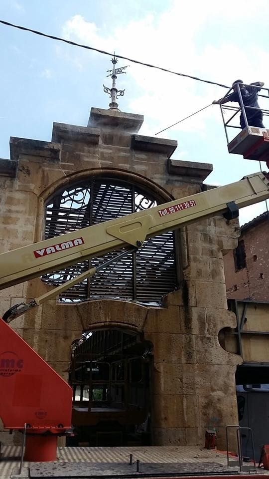 Un trabajador, subido a una plataforma elevadora hidráulica, trabaja en la fachada de piedra de un arco histórico coronado por una veleta.