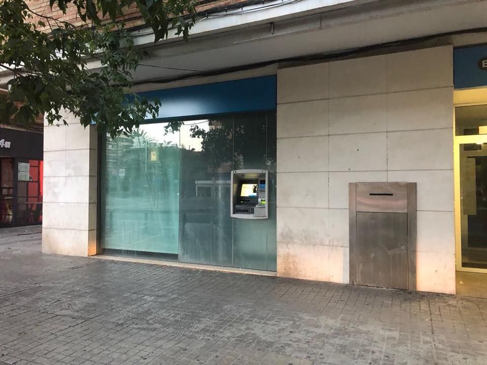Vista a nivel de calle del exterior de un edificio bancario, con una ventana de cristal, un cajero automático y una caja de seguridad metálica.